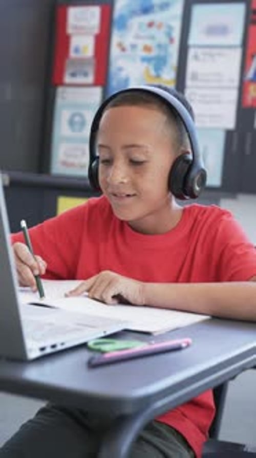 Vertical video: In school, child wearing headphones writing in notebook at desk with laptop