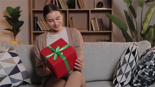 Smiling Caucasian Woman Sitting on Sofa Holding Red Gift Box with Green Ribbon Happy Female Looking