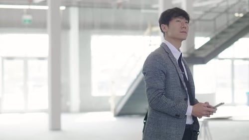 Mature asian property owner visualizing expansion in vacant warehouse with smart phone