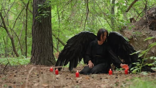 Dark Winged Woman Practicing Spiritism with Red Candles in Forest