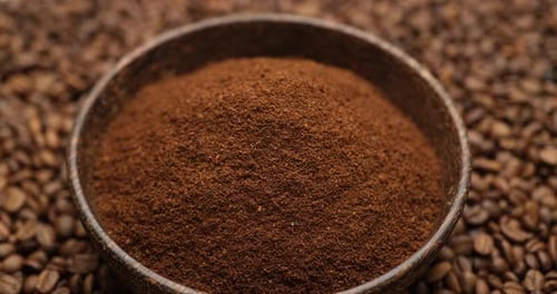 Holzschale voller gemahlener Kaffeebohnen auf rotierendem Hintergrund aus frisch gerösteten Kaffeebohnen