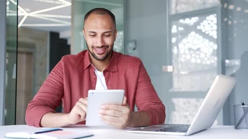 Un hombre de negocios sonriente está usando una tableta digital mientras está sentado en su lugar de trabajo en una oficina comercial moderna.