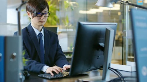 In the Office East Asian Businessman works on a Desktop Personal Computer. His Colleague's Computer