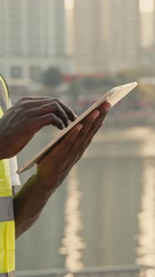 Crop View of African American Builder Holds Smart Tablet Working on Screen Standing Against Urban