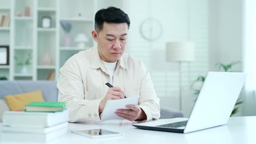 Concentrated asian man watching video call lesson or training using laptop while sitting in home