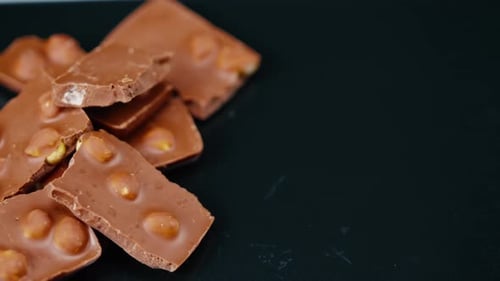 Nahaufnahme auf schwarzblauem Hintergrund auf einem schwarzen Teller liegt Milchschokolade mit eingebrochenen Haselnüssen