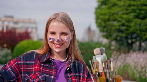 A young girl artist paints a picture with a brush on a canvas that stands on an easel view behind