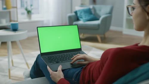 Young Woman at Home Works on a Laptop Computer with Green Mock-up Screen. She's Sitting On a Couch