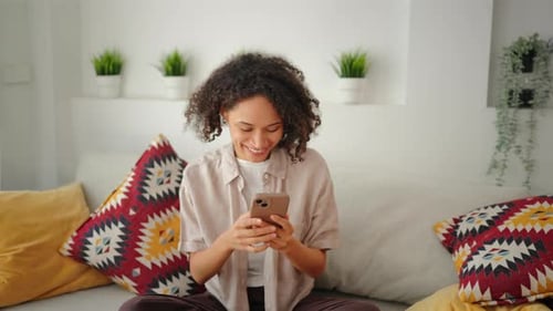 Smiling Young Woman Uses Phone on Sofa Indoors
