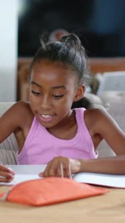 Vertical video: Leaning girl in pink tank doing work on pages at wooden table, reaching pencil case