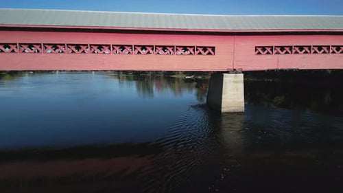 An aerial drone view underneath a red and gray covered walking bridge across a river during the fall