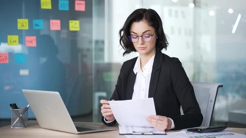 Professional female accountant analyzes financial documents in office setting. Woman uses laptop