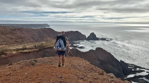Male backpacker walks to edge of tall ocean cliff to enjoy the view