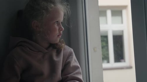 Sad girl looks out the window through the glass on a cloudy, gray day.