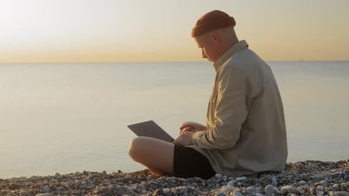 A Young Man Sitting on the Beach and Working on a Laptop