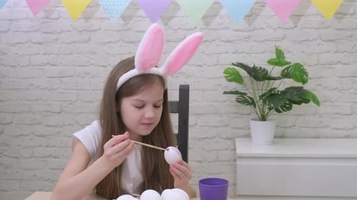 Kleines Mädchen mit Hasenohren bemalt Eier für Ostern zu Hause