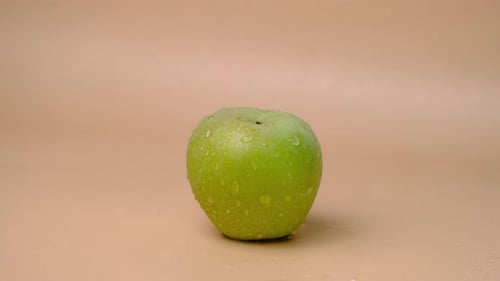 Slow motion shot of water splashing on fresh green apple on beige background.