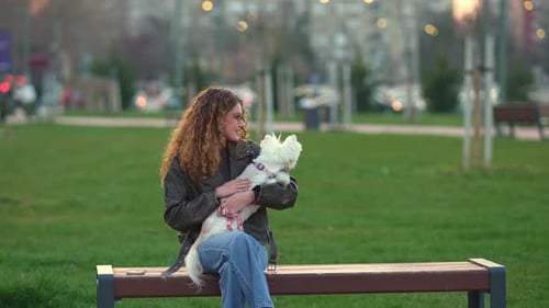 Relaxed young woman sitting on a park bench with her small white dog, enjoying a peaceful moment out