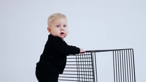 Lovely Caucasian baby with blond hair wearing black suit. Kid stands holding by the cage.