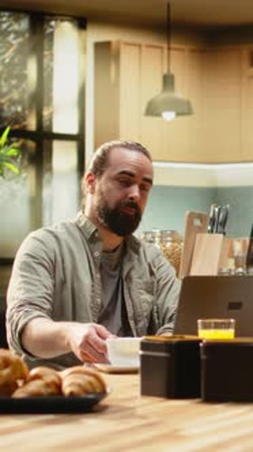 Man with Ponytail Talks on Laptop at Kitchen Table