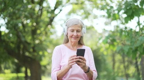 Senior active gray haired woman in headphones uses a smartphone while standing in an urban city park