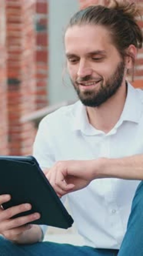 Handsome Caucasian Man in White Shirt with Beard Sitting on Stairs and Working on Computer Tablet at