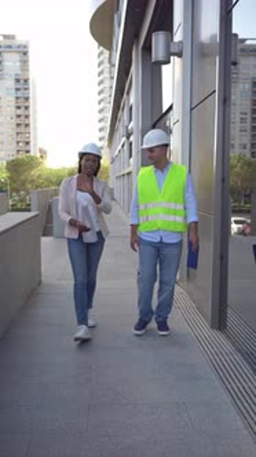 Two diverse architects and engineers in safety helmets and vests walking and talking outside office