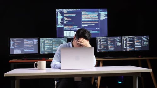 Programador masculino asiático que tiene dolor de cabeza mientras escribe código con una computadora portátil con varios monitores