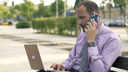 Young Businessman With Laptop Talking On Cellphone Sitting On Bench In City