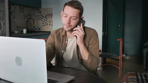 Man Talking on Smartphone While Working on Laptop at Home Office Table Male Digital Nomad on Remote