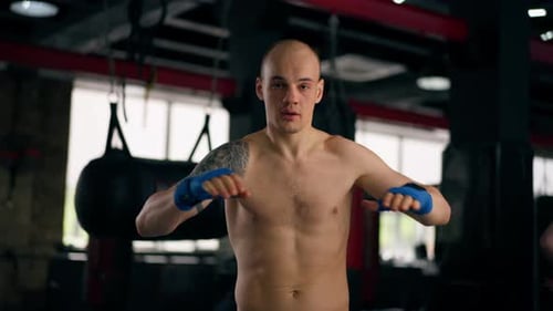Closeup in a Boxing Fight Club Bald Young Boxer with Tattoos in Blue Bandages Trains Punches Shadow