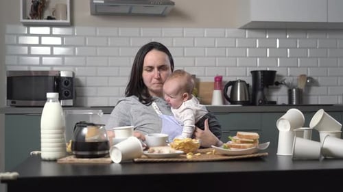 Tired Mother Holding Baby Drinking Coffee at Messy Table
