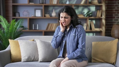 Upset young woman suffering from toothache while sitting on sofa in living room at home. The female