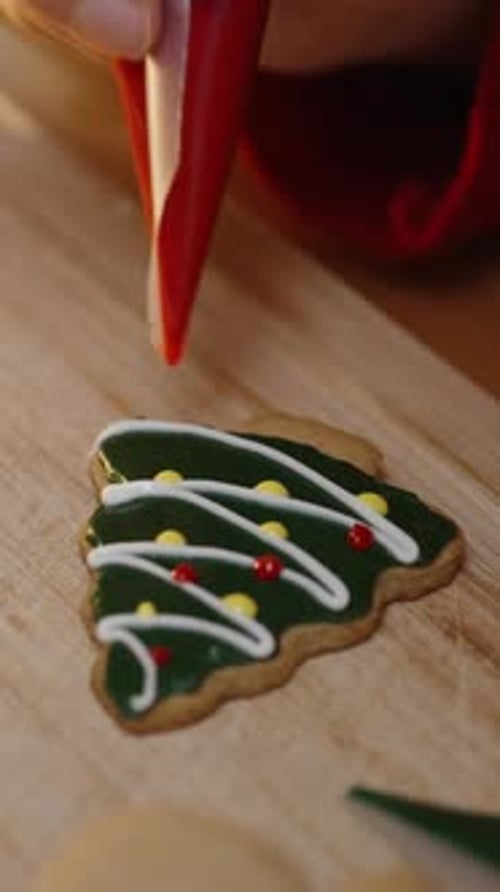 Vertical video. Carefully Using Red Icing, She Adds Festive Touches to a Green Christmas Tree Cookie