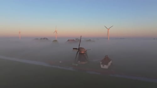 Sunrise drone shot over misty meadow with windmill, houses, and barns