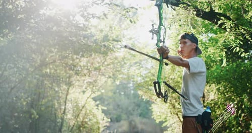 Slow motion of young man with professional equipment is practising archery with a bow in a woods i