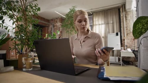 Female Employee Using Laptop and Checking Smartphone at Desk in Modern Office