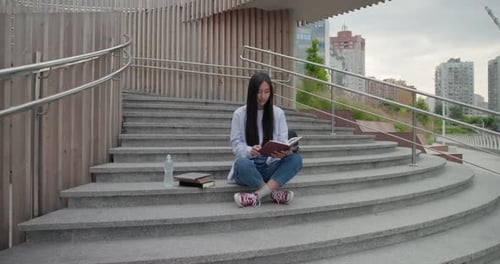 Education in University College Concept Young Lady Reading Notebook While Sitting Outdoors on Steps