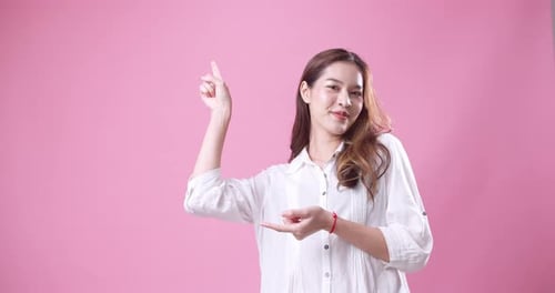Beautiful young Asian woman pointing to the right side. Studio recording with a pink background.