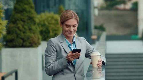 Businesswoman Smiling While Checking Phone and Holding Coffee Outside Office Building
