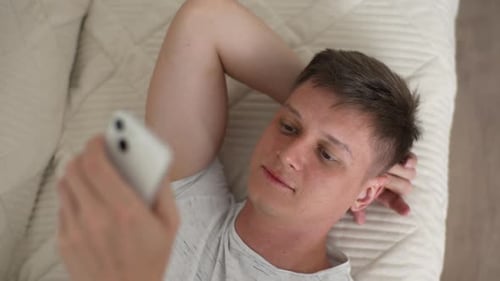 Closeup Top View Portrait of Young Carefree Relaxed Man Lying on Sofa and Holding Phone Chatting