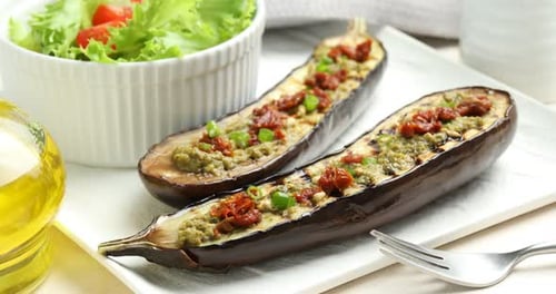 Slices of grilled eggplant with sun dried tomatoes, pesto and salad on white table, closeup