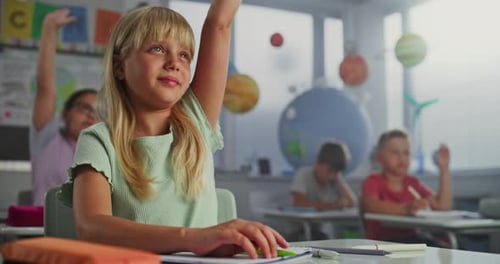 Primary School Girl Sitting at Desk Raising Hand and Reading Correct Answer From Notebook