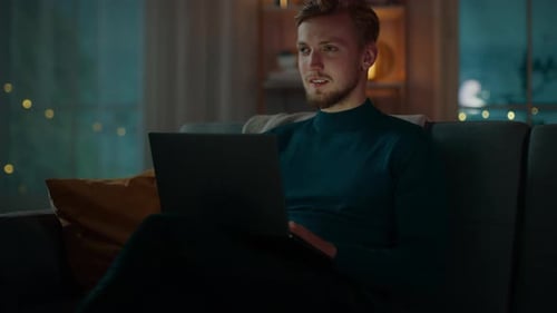 À noite, um jovem bonito em casa sentado em um sofá trabalha em um laptop.
