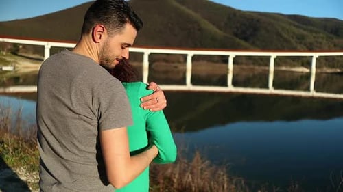 Caucasian guy hugs and caresses a red-haired girl dressed in green by the lake at sunset, love conce