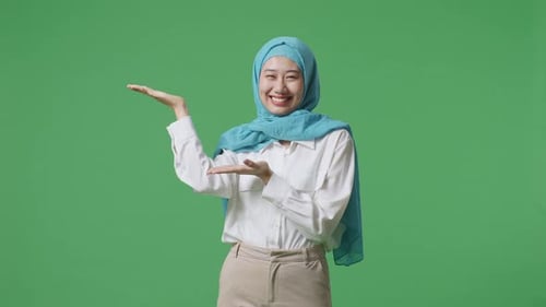Asian Muslim Woman Smiling And Pointing To Side While Standing In The Green Screen Background