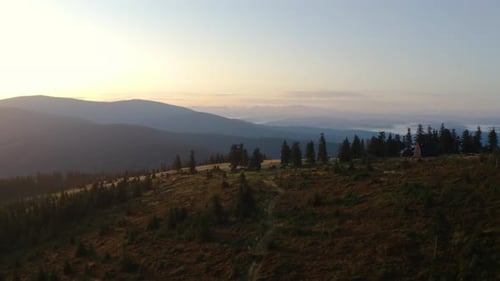 Drone flight forward above forested mountain's top during sunrise, foggy lowland in the distance