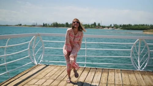 Pretty young woman dances on wooden pier near shore line of picturesque sea with blue sky.
