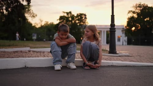 Das glückliche kleine blonde Mädchen in einem rosa T-Shirt sitzt auf dem Bordstein und kommuniziert mit ihrem Bruder, der