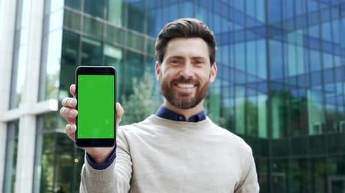 Confident smiling male entrepreneur holding phone with green screen for customization standing